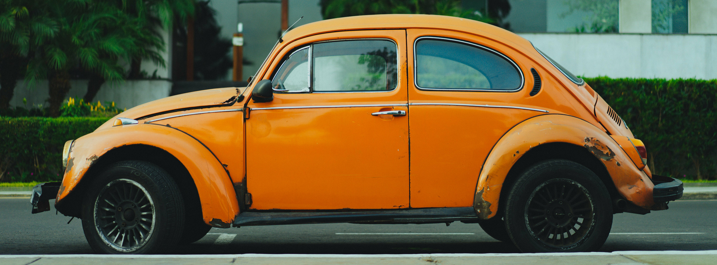 Ein oranger VW Käfer mit offensichtlichen Rostfelcken und Dellen. Im Hintergrund befindet sich ein Haus