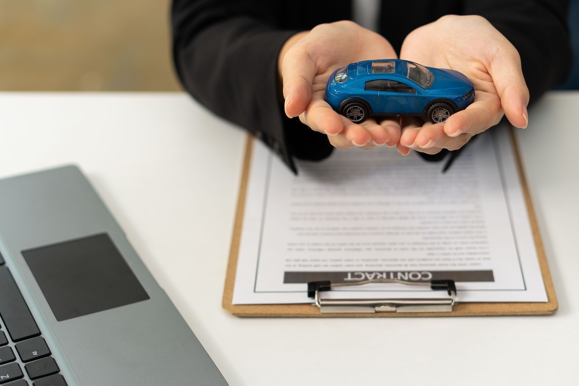 Ein Mann hällt ein Spielzeugauto über einen schriftlichen KFZ-Vertrag vor ihm steht ein Laptop 